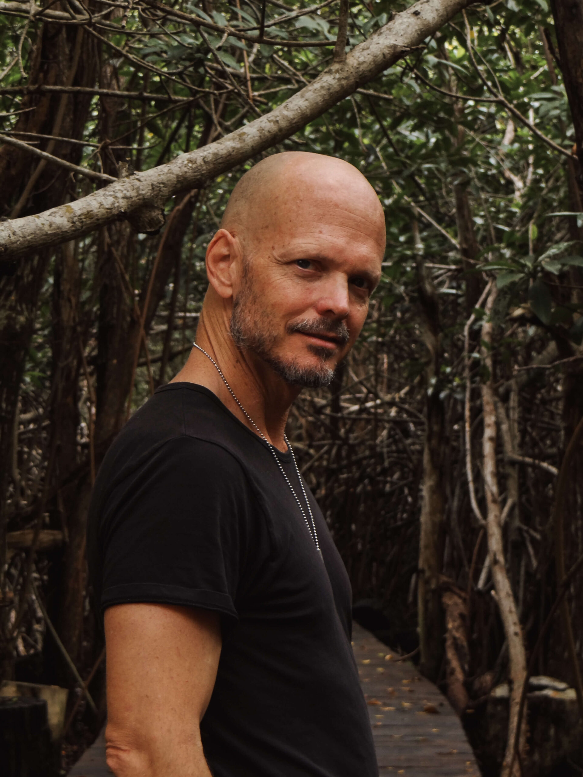 Brian Begin A person with a shaved head and beard in a black shirt stands on a wooden path in a dense forest setting, exuding an aura of spirituality and personal growth while looking at the camera. Brian Begin Podcast