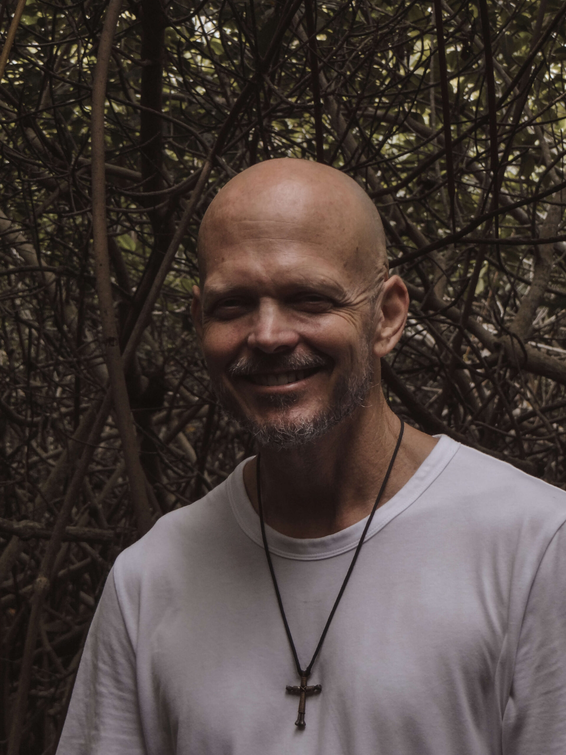 Brian Begin A man with a bald head and beard, wearing a white shirt and cross necklace, smiles warmly while standing amidst dense, intertwining branches, radiating an aura of spirituality and personal growth. Brian Begin Podcast