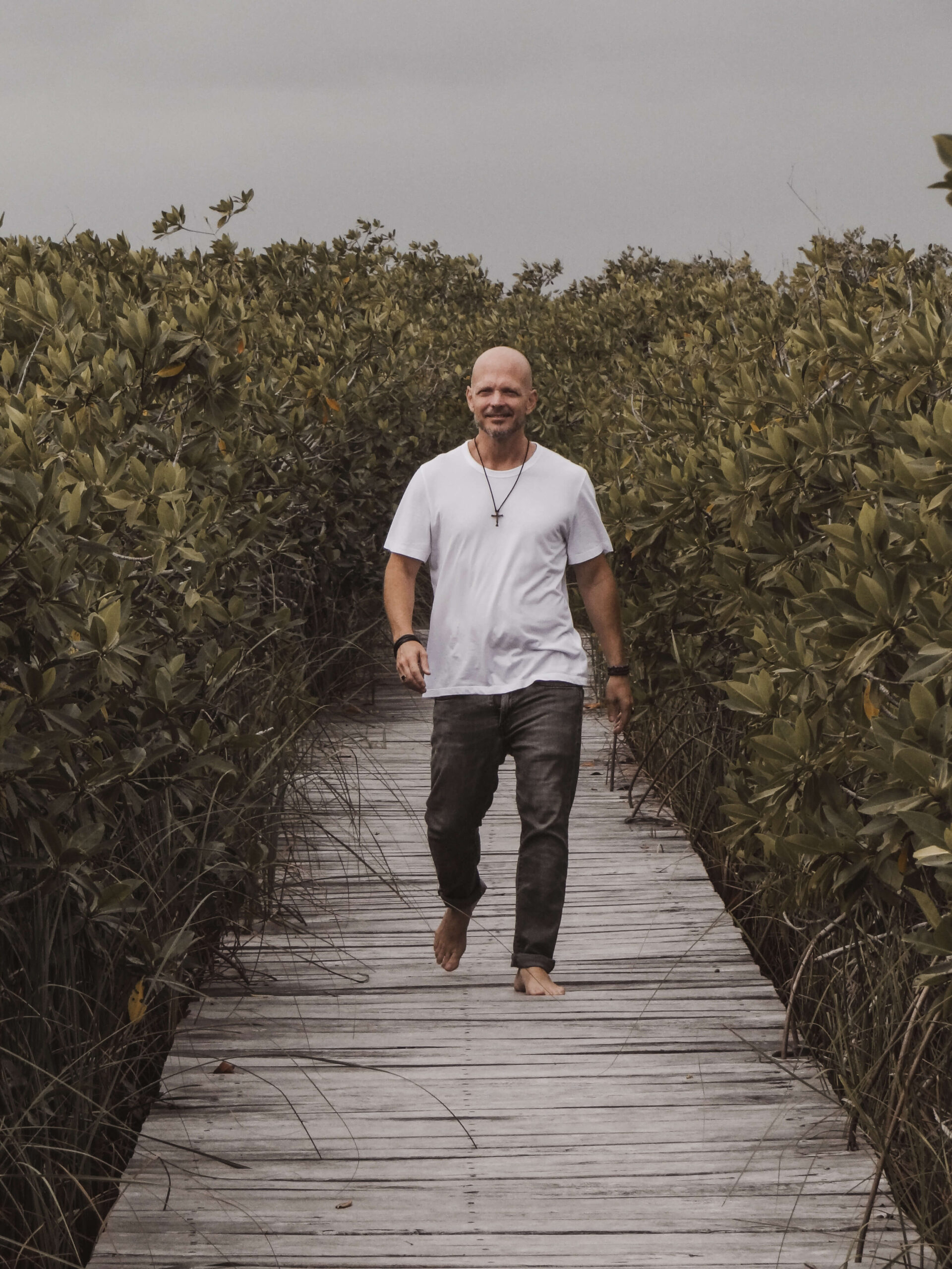 Brian Begin Bald man in a white t-shirt and jeans walks barefoot on a wooden path through dense green foliage, embracing spirituality and personal growth with each step. Brian Begin Podcast