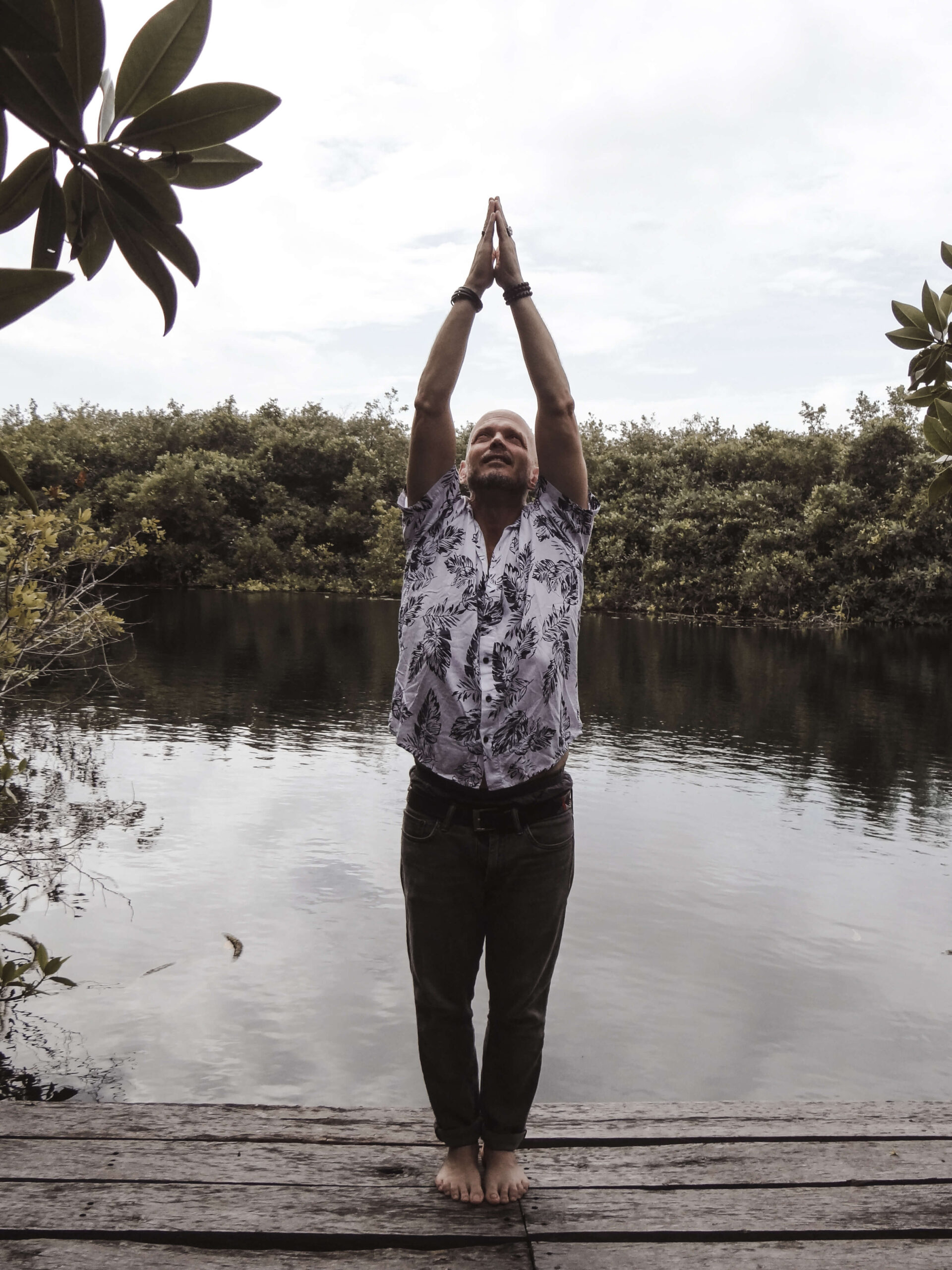 Brian Begin Barefoot on a wooden platform by the lake, surrounded by trees, a person embraces spirituality with arms raised in a yoga pose. This serene moment of connection reflects personal growth akin to insights from the Brian Begin Podcast. Brian Begin Podcast
