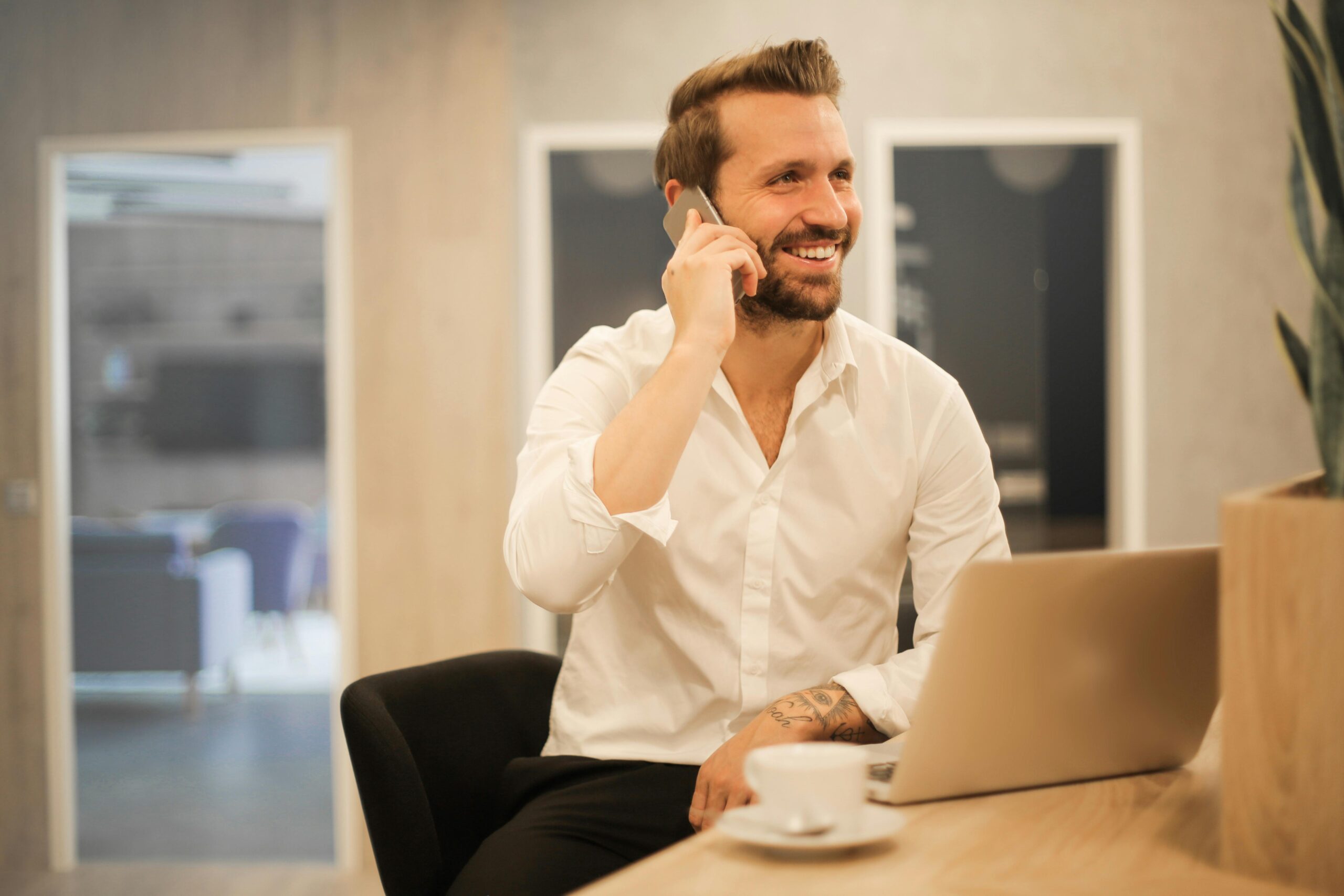 Brian Begin Man in a white shirt sits at a desk with a laptop and coffee cup, smiling while talking on a phone in a modern office setting. Brian Begin Podcast