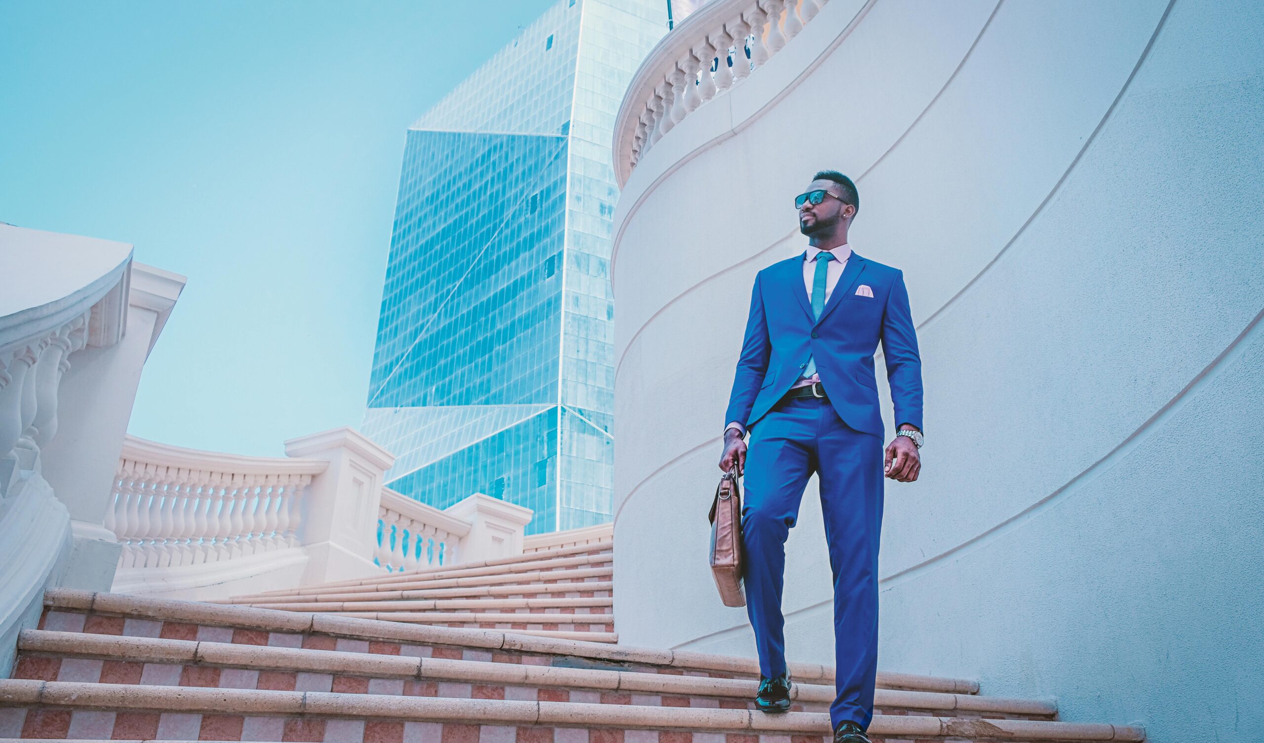 Brian Begin A man in a blue suit walks up outdoor stairs, holding a briefcase, with modern glass buildings and ornate railings in the background. Brian Begin Podcast