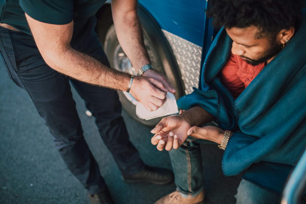 Brian Begin A person sitting on the ground receives first aid from another individual, who is cleaning a wound on the seated person's wrist. Brian Begin Podcast