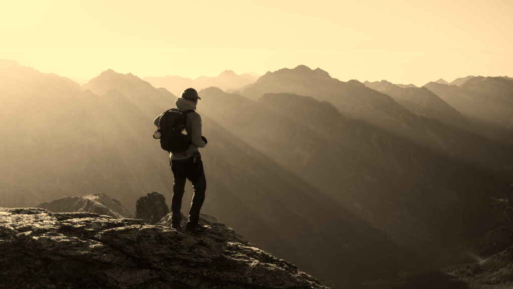 Brian Begin A person with a backpack stands on a rocky mountain ledge, looking out at a range of mountains under sunlight streaming through mist. Brian Begin Podcast