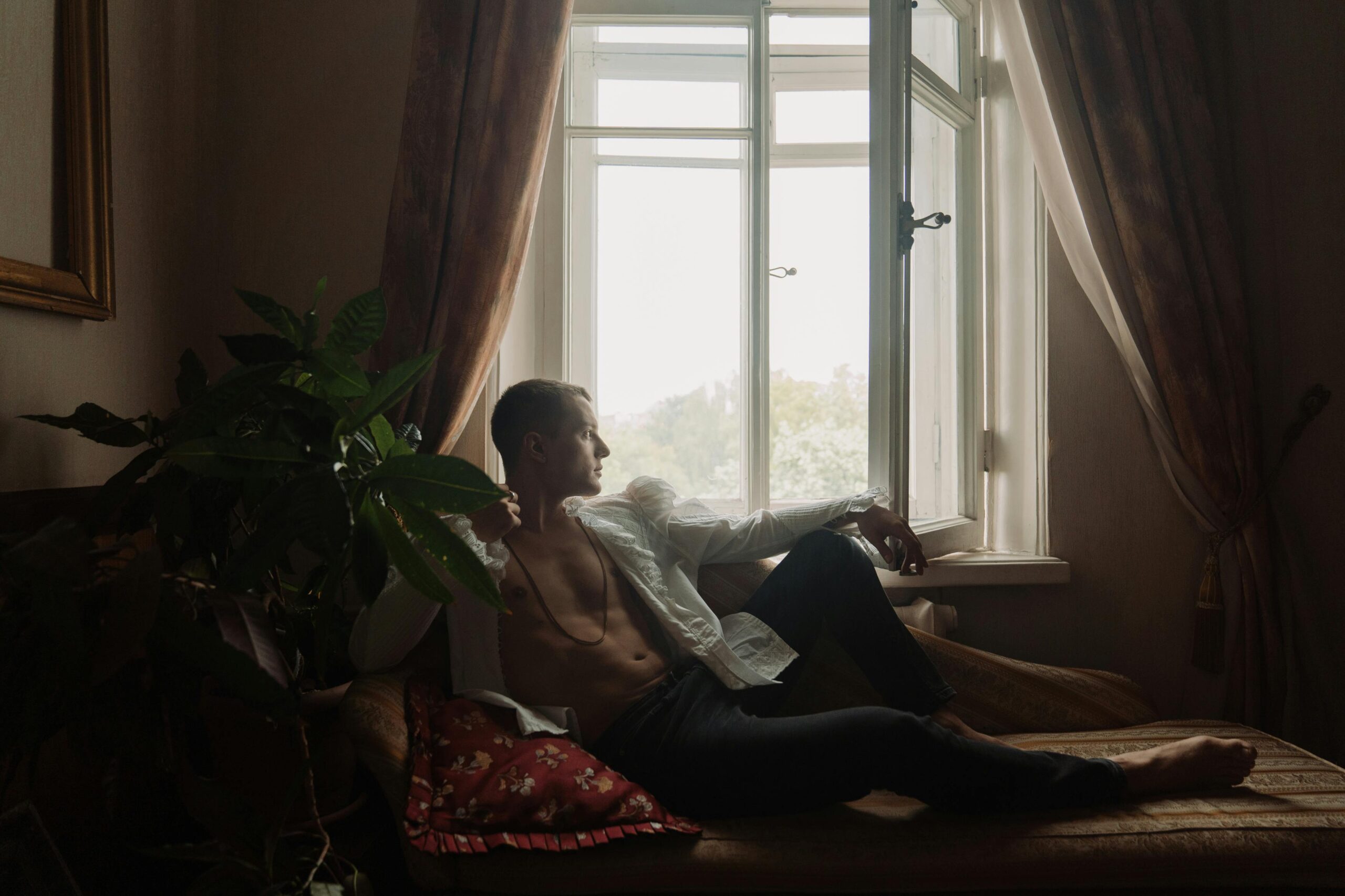 Brian Begin A man with an open shirt sits on a couch by a window, looking outside; sunlight filters through sheer curtains and a potted plant is nearby. Brian Begin Podcast