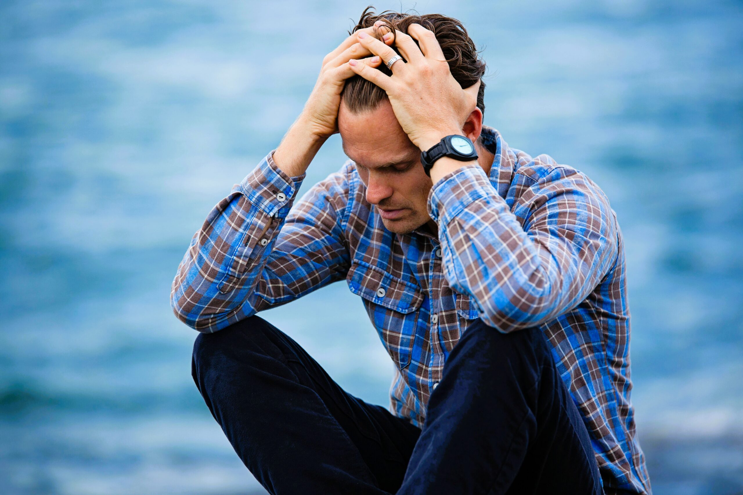 Brian Begin A man in a plaid shirt sits with knees up, holding his head in both hands, appearing stressed, with a blurred body of water in the background. Brian Begin Podcast