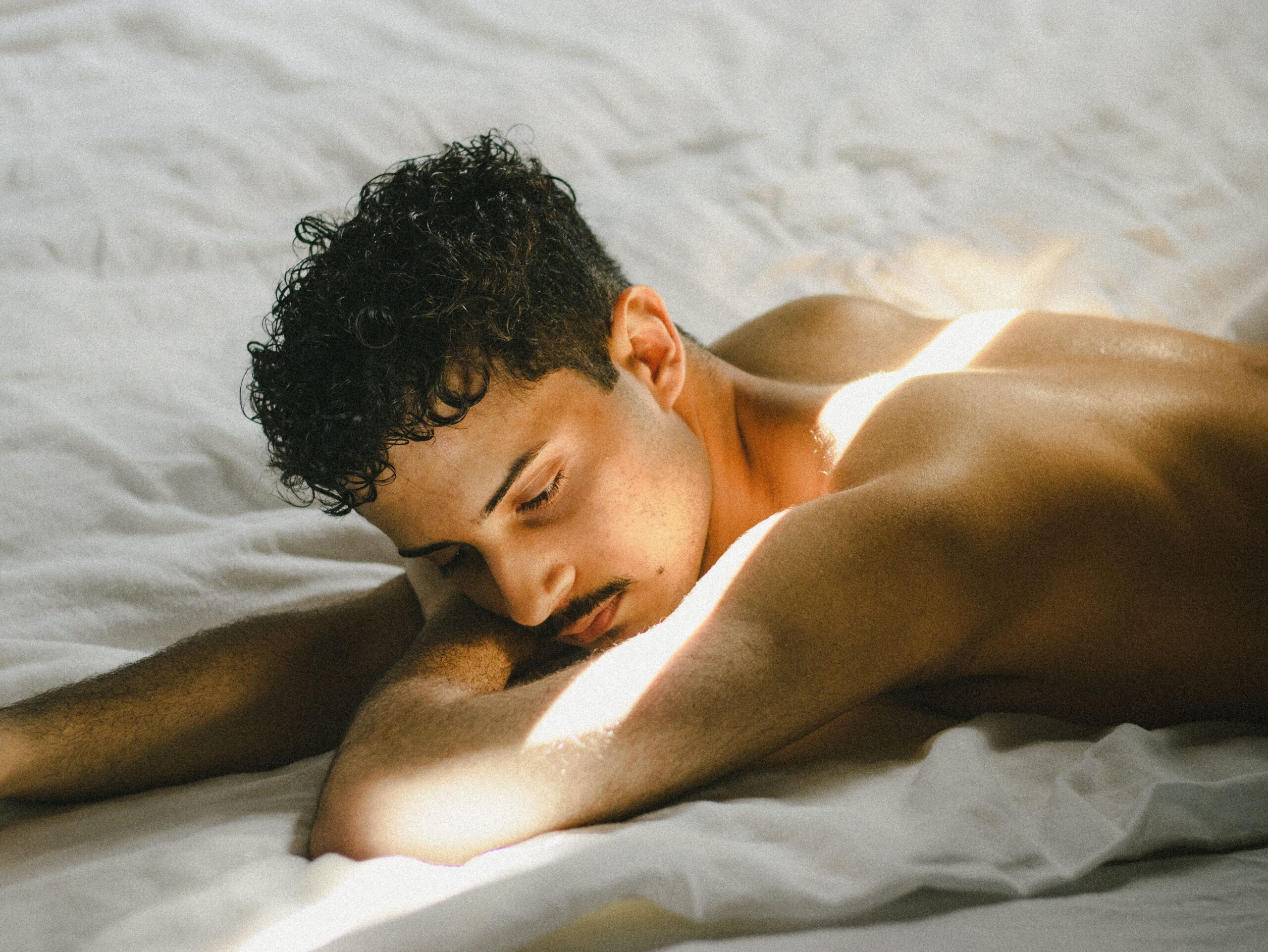Brian Begin A person with short curly hair lies shirtless on a bed, resting their head on their arm, with sunlight streaming across their body and white bedding. Brian Begin Podcast