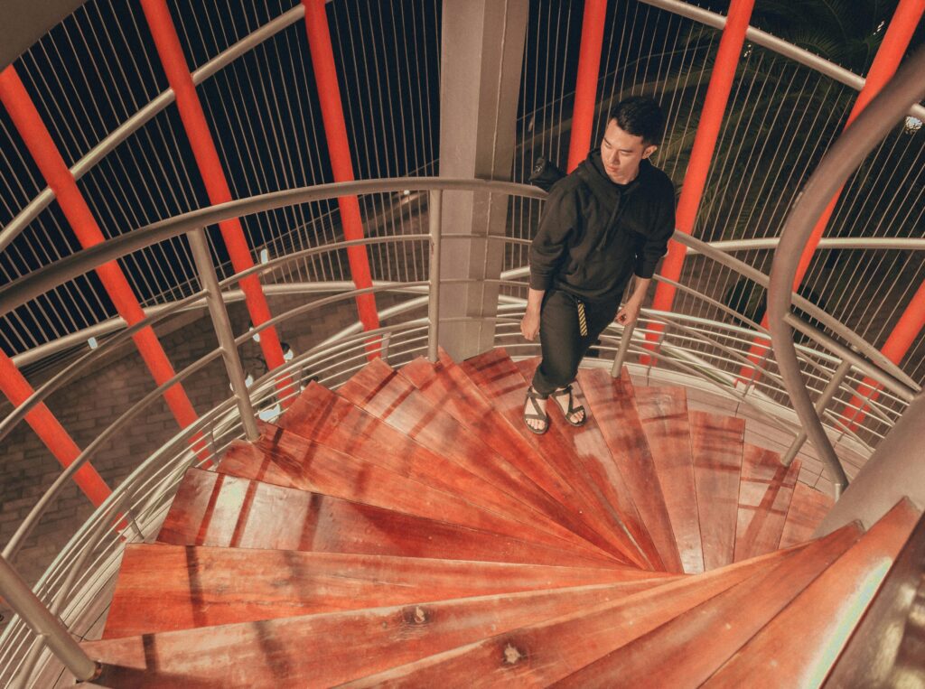 A person in dark clothing walks up a spiral wooden staircase with red vertical bars, viewed from above at night.