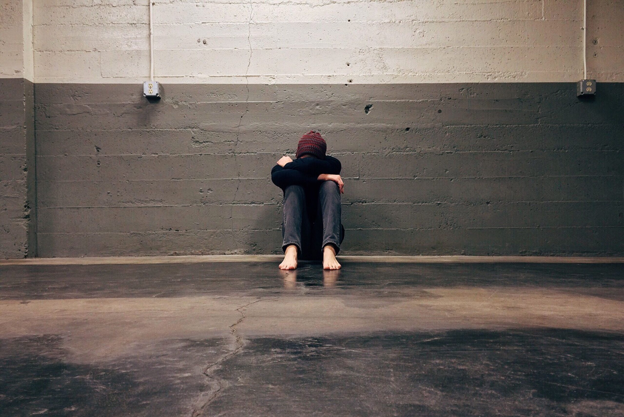 Brian Begin A barefoot person sits alone on the floor in an empty room, knees drawn to chest, arms wrapped around legs, head down against arms, against a gray concrete wall. Brian Begin Podcast