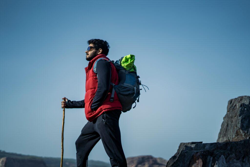A person wearing a red vest and backpack stands on rocky terrain, holding a walking stick and looking into the distance under a clear sky